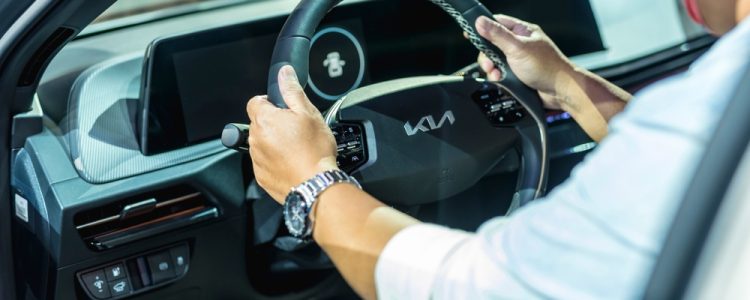 A person gripping the steering wheel of a Kia car, showing the dashboard and modern controls inside the vehicle