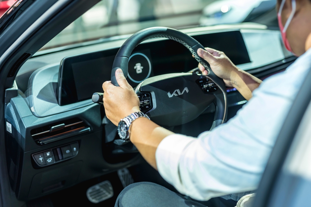 A person gripping the steering wheel of a Kia car, showing the dashboard and modern controls inside the vehicle