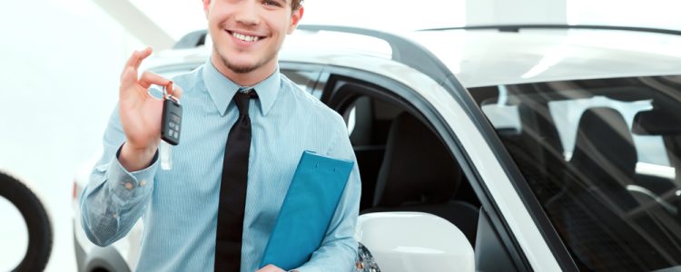 A smiling car salesman in a light blue shirt and black tie holding car keys and a clipboard, standing in front of a white car in a showroom