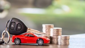 A toy red car with car keys resting on top, positioned next to several stacks of coins, symbolizing car expenses or financing