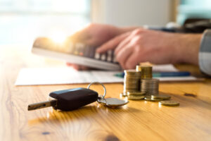 Car key and stacked coins on a table with a person using a calculator in the background, suggesting financial planning or budgeting