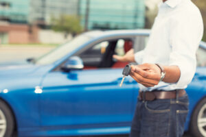 Man standing next to a blue car holding out a car key, suggesting sale, rental, or transfer of vehicle ownership