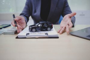 person gestures toward a miniature black SUV on a contract, with stacks of cash, a pen, and a laptop on the desk, suggesting car purchase or financing