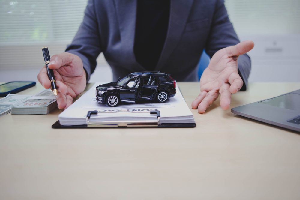 person gestures toward a miniature black SUV on a contract, with stacks of cash, a pen, and a laptop on the desk, suggesting car purchase or financing