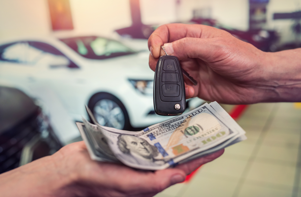 Person handing over car keys in exchange for a stack of hundred-dollar bills at a car dealership