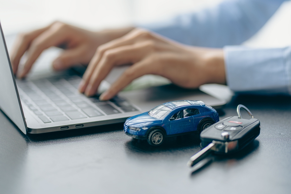 Person typing on a laptop with a toy car and car keys on the desk, suggesting online car shopping or vehicle-related research