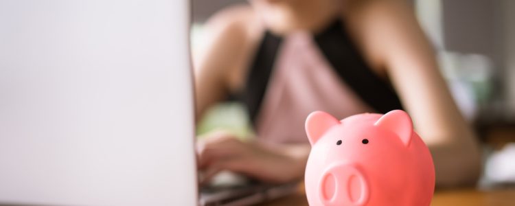 Pink piggy bank sits on a wooden desk in the foreground, with a blurred woman using a laptop in the background