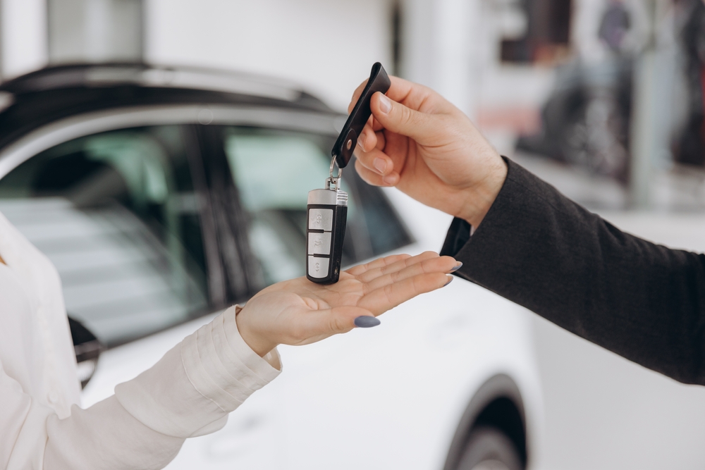 Close-up of a person handing car keys to another person, with a white car in the background