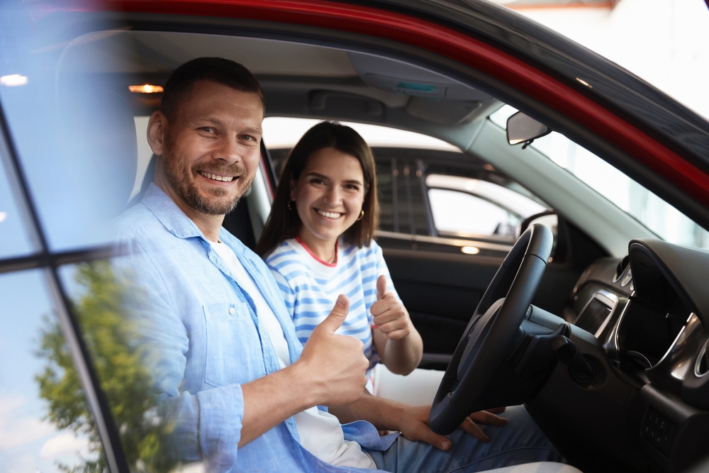 Smiling man and woman sitting in a car giving thumbs up, suggesting satisfaction with a new vehicle or test drive experience