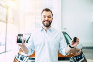 Smiling man standing in front of a car, holding a smartphone in one hand and a key fob in the other, suggesting a new car purchase or digital car service