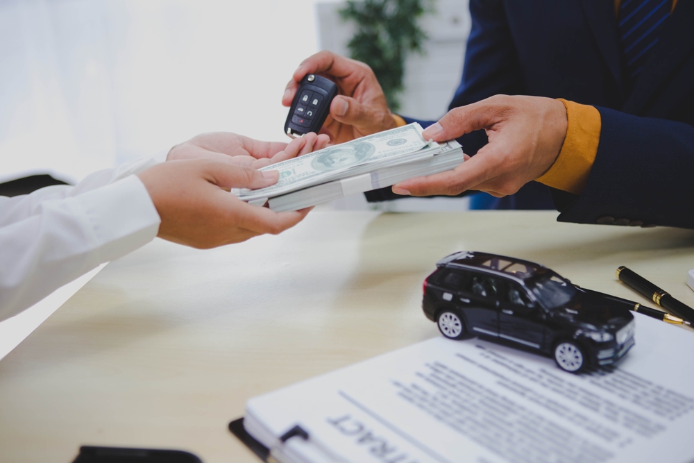 Two people exchange car keys and cash at a desk with a toy SUV and a contract, symbolizing a car sale or purchase