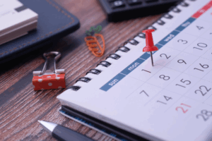 Close-up of a desk calendar with a red pushpin marking Monday the 1st, surrounded by office supplies