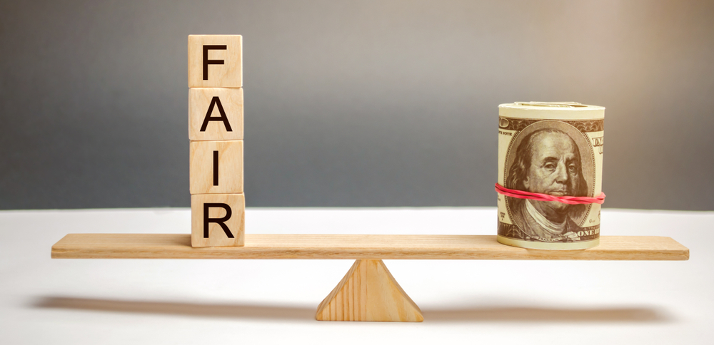 Wooden blocks spelling “FAIR” balance a roll of U.S. hundred-dollar bills on a wooden seesaw, symbolizing fairness in financial matters